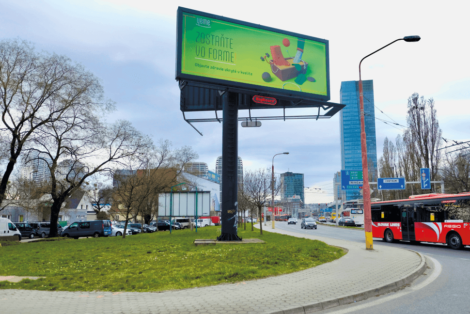 Záber na predchádzajúcu podobu Mlynských nív s veľkou reklamnou plochou na stĺpe na trávniku vedľa chodníka a cesty.